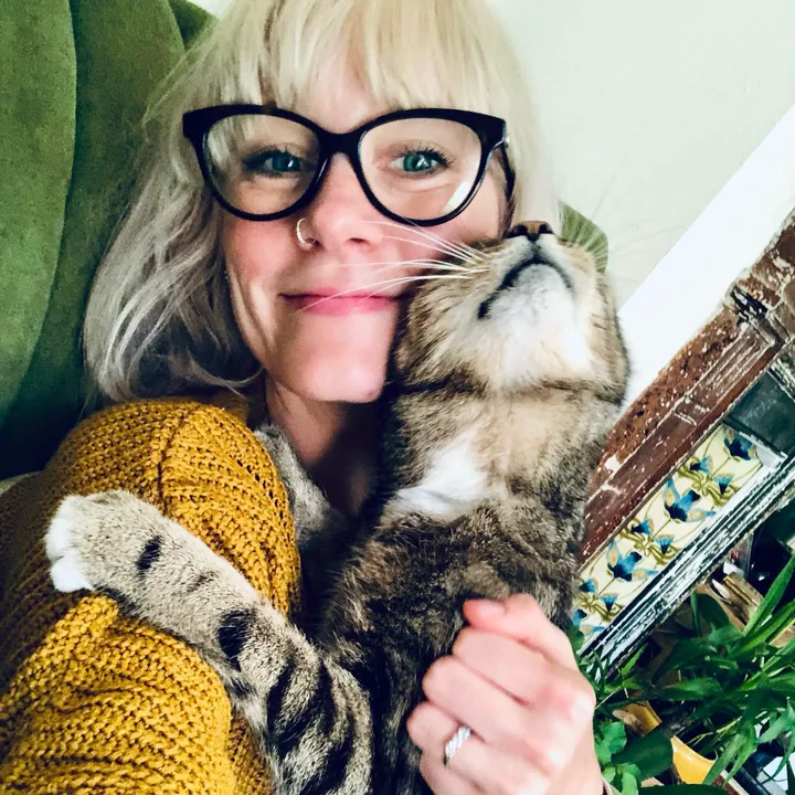 Milly smiling with a tabby cat nuzzled against her cheek